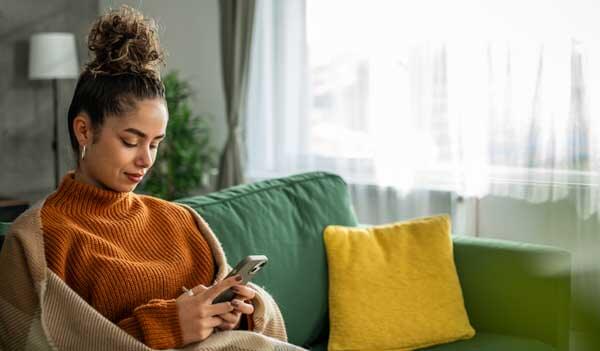 Woman on sofa wrapped in blanket using phone